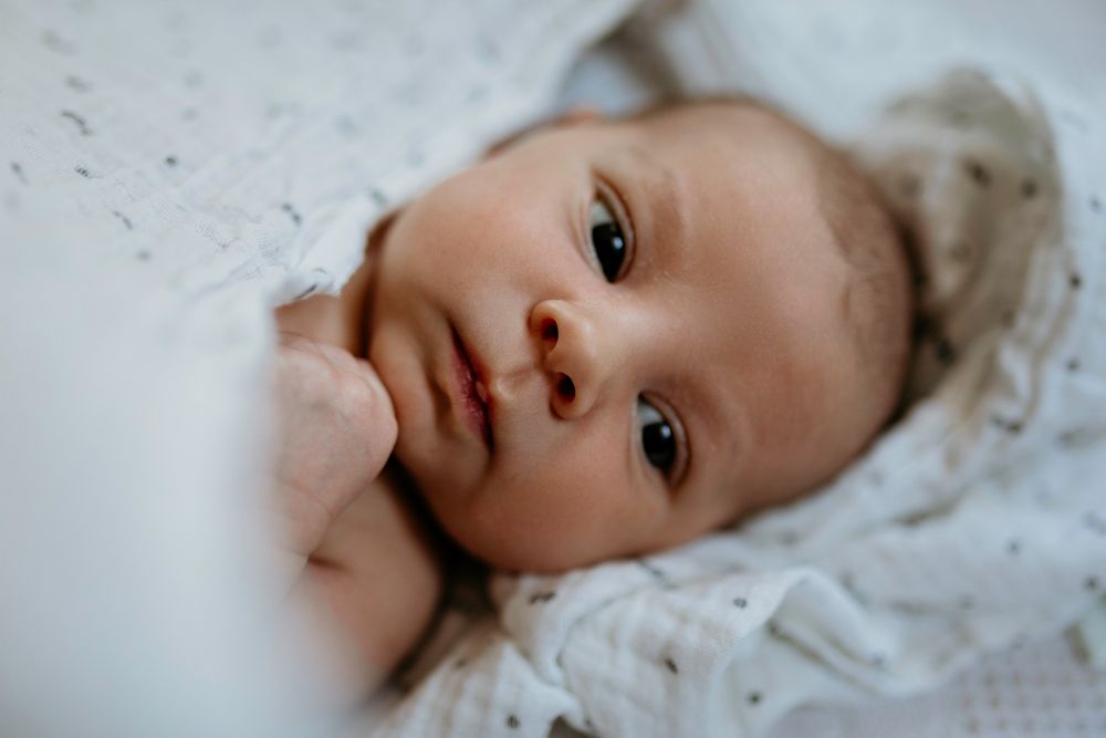 A sweet new baby with bright eyes lies surrounded by soft white fabric in a gentle close-up portrait.