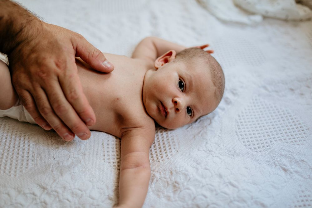 A newborn baby lies on white textured bedding while a parent's hand gently rests against their back.