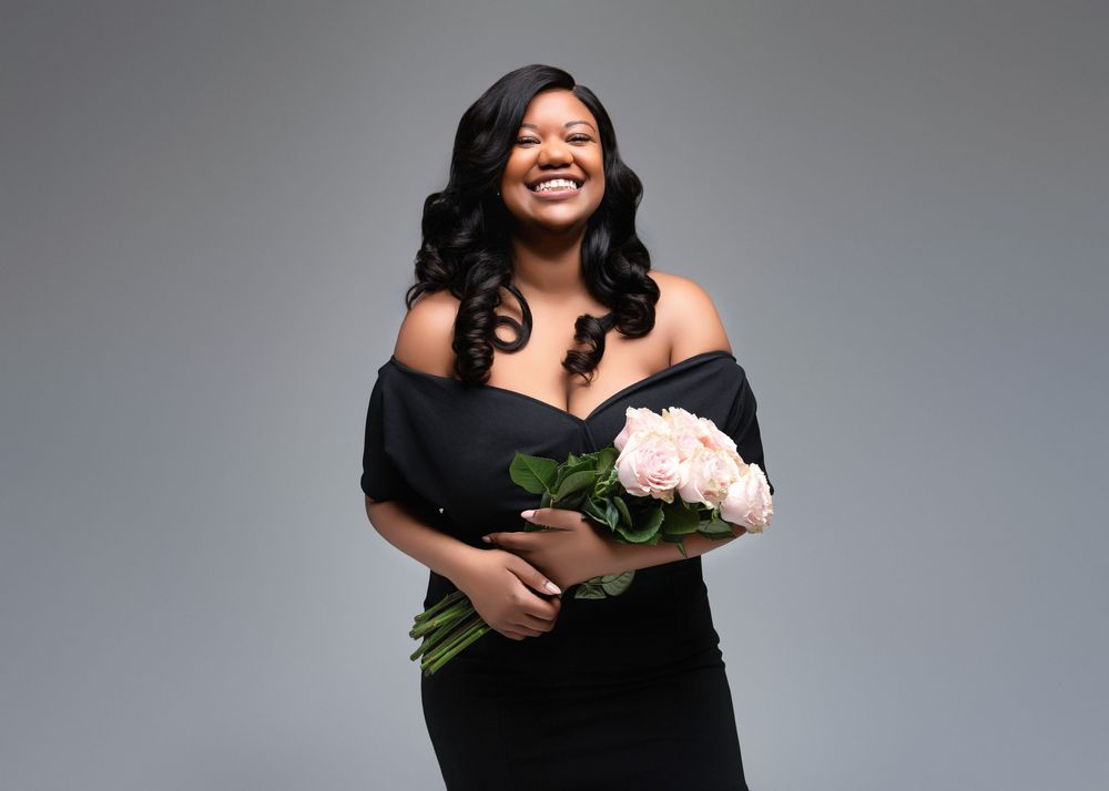 A person in an elegant black off-shoulder dress holds pink roses while posing against a gray backdrop.