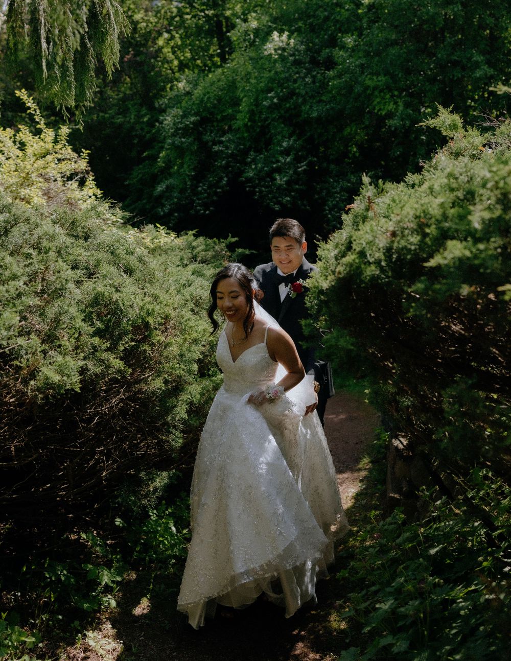 A couple walks along a garden path surrounded by lush greenery and willow trees on their wedding day.