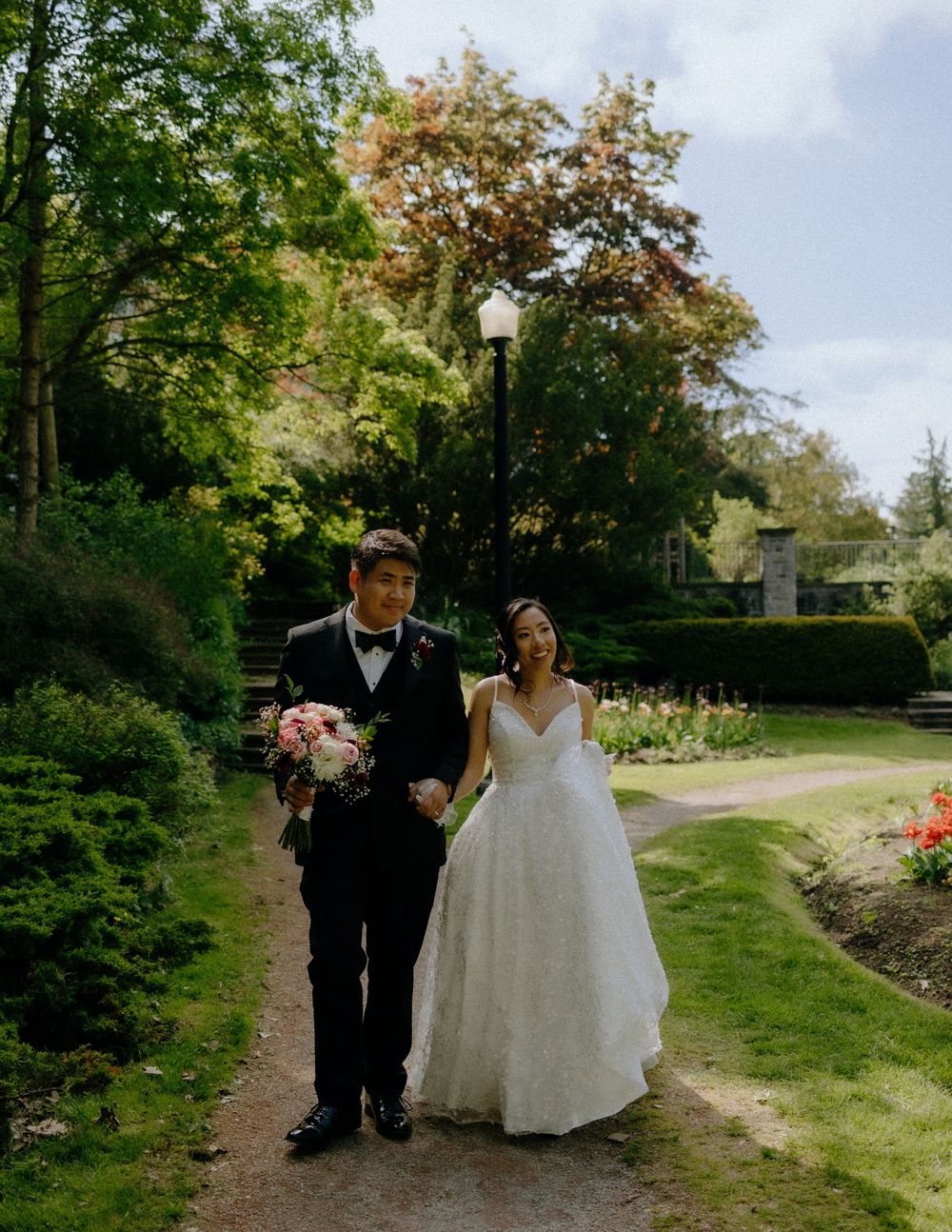 A couple walks together along a garden path surrounded by lush greenery and flowers on their wedding day.
