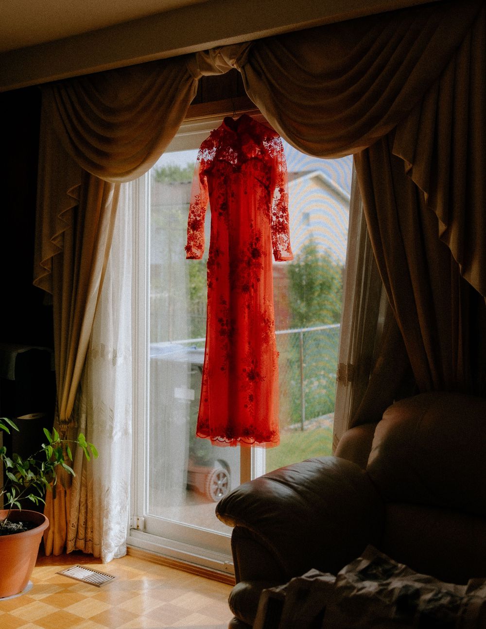 A vibrant red traditional Chinese qipao dress with lace details hangs elegantly against a sunlit window with brown curtains.