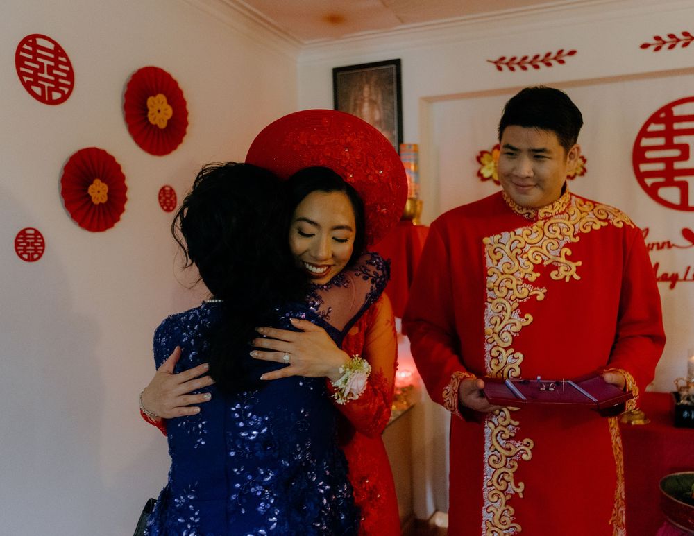 Wedding guests embracing in a room decorated with traditional Chinese double happiness symbols and red decor.