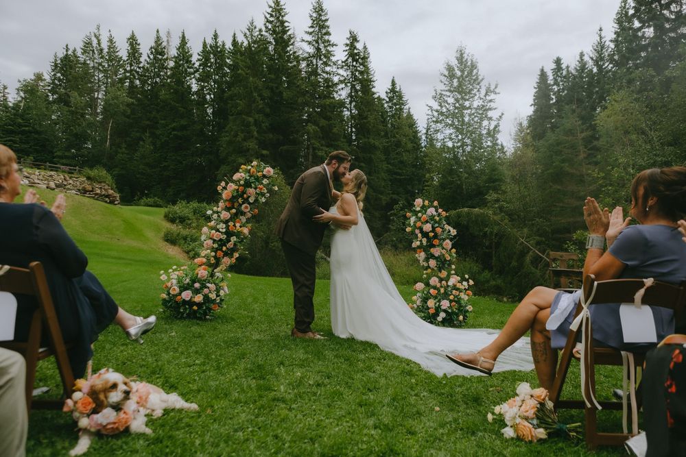 A romantic outdoor wedding ceremony with floral arch and guests seated on a lush green lawn surrounded by evergreen trees.