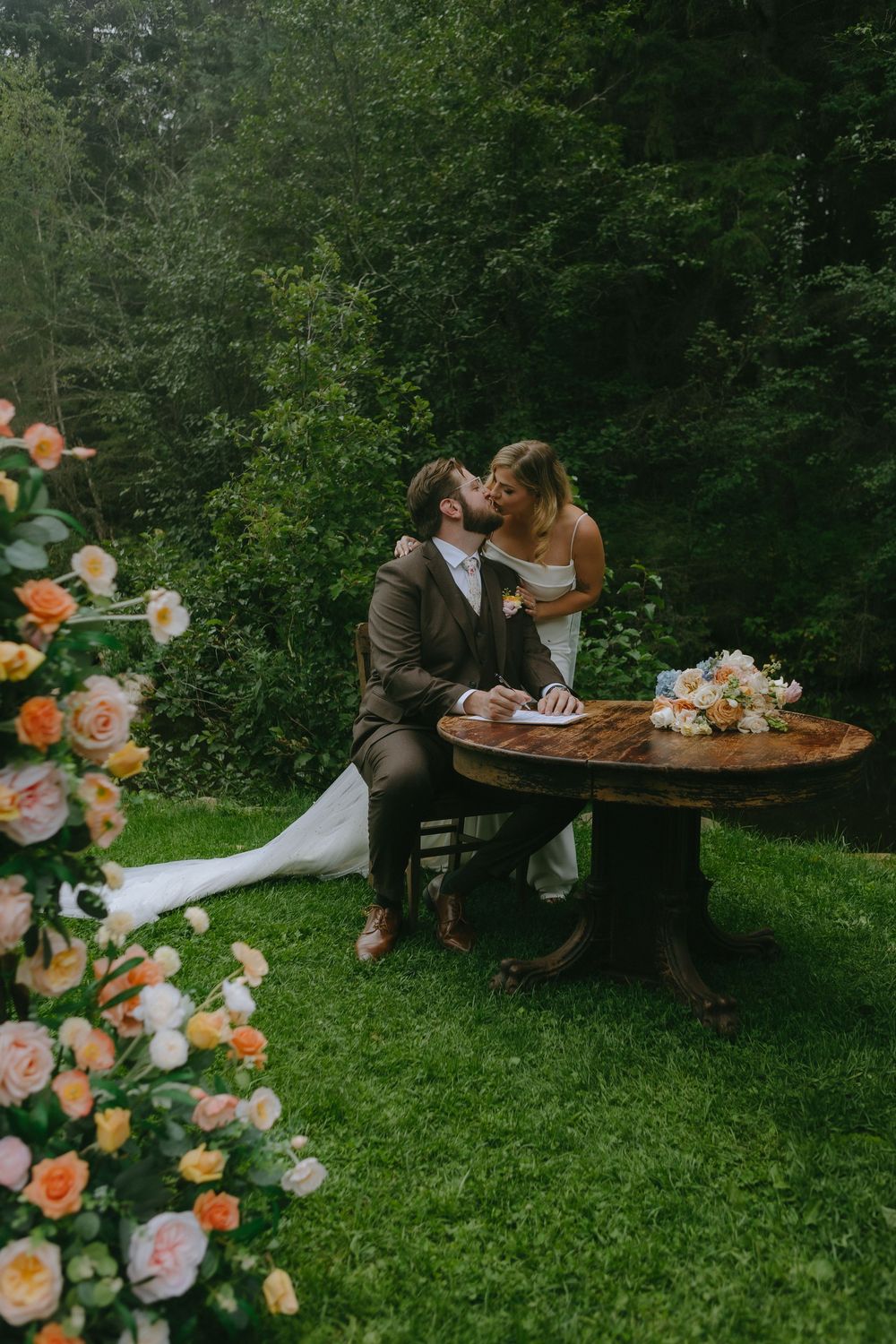 A couple shares an intimate moment at a rustic wooden table surrounded by peach roses and greenery.