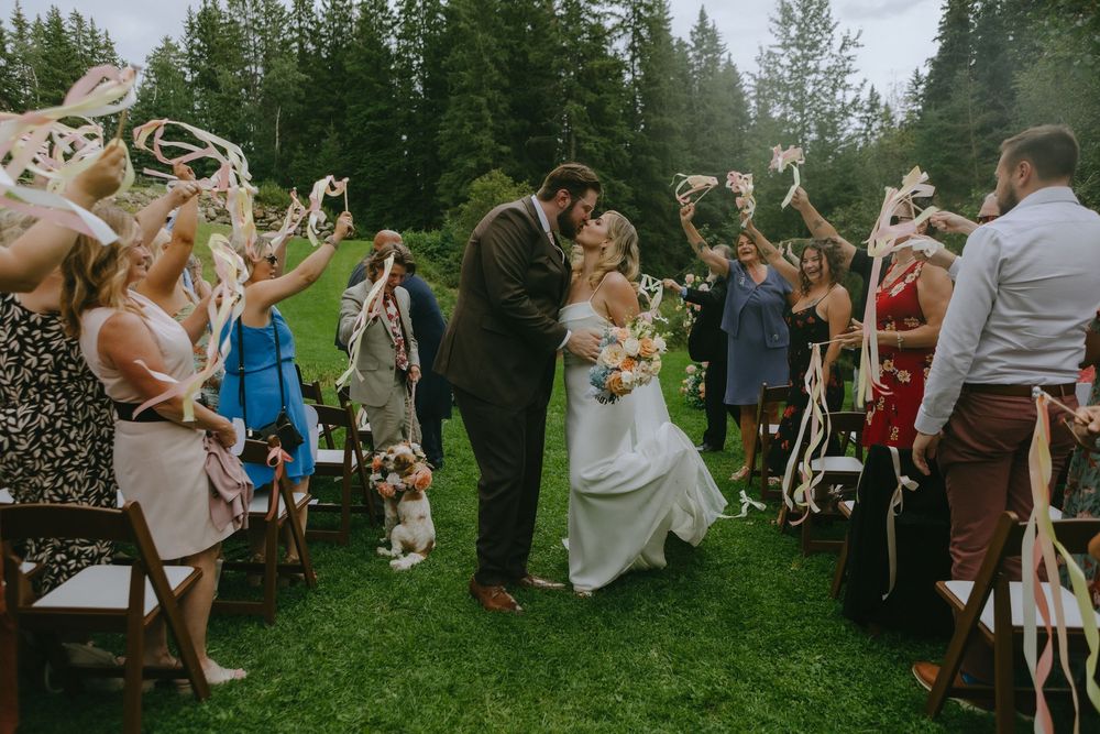Wedding guests toss confetti over a couple dancing on a lush green lawn surrounded by pine trees.