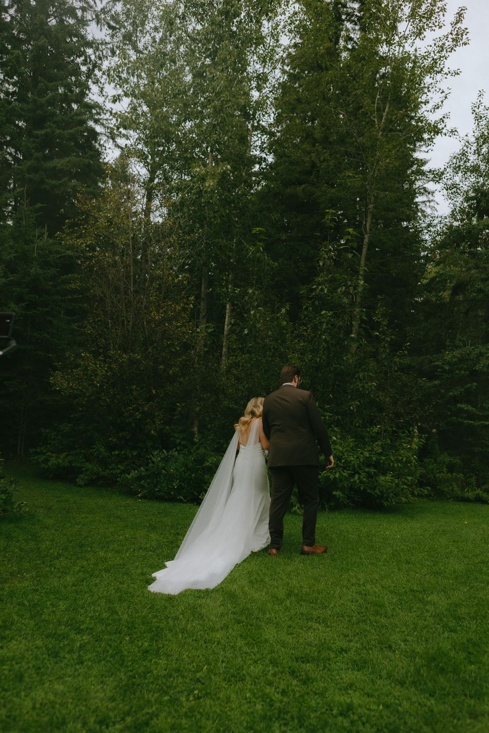 A couple in wedding attire walking through a lush forest setting with tall evergreen trees and green grass.