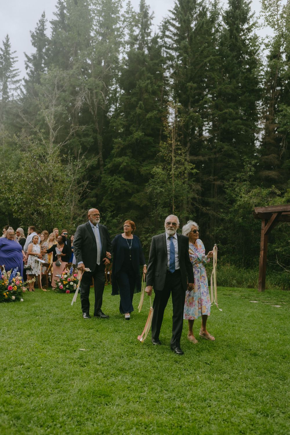 Outdoor wedding ceremony in a green forest setting with attendees gathered on grass and pine trees in background.