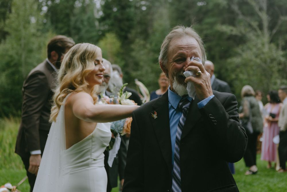 Joyful wedding celebration with laughter and embraces on a grassy lawn with forest backdrop.