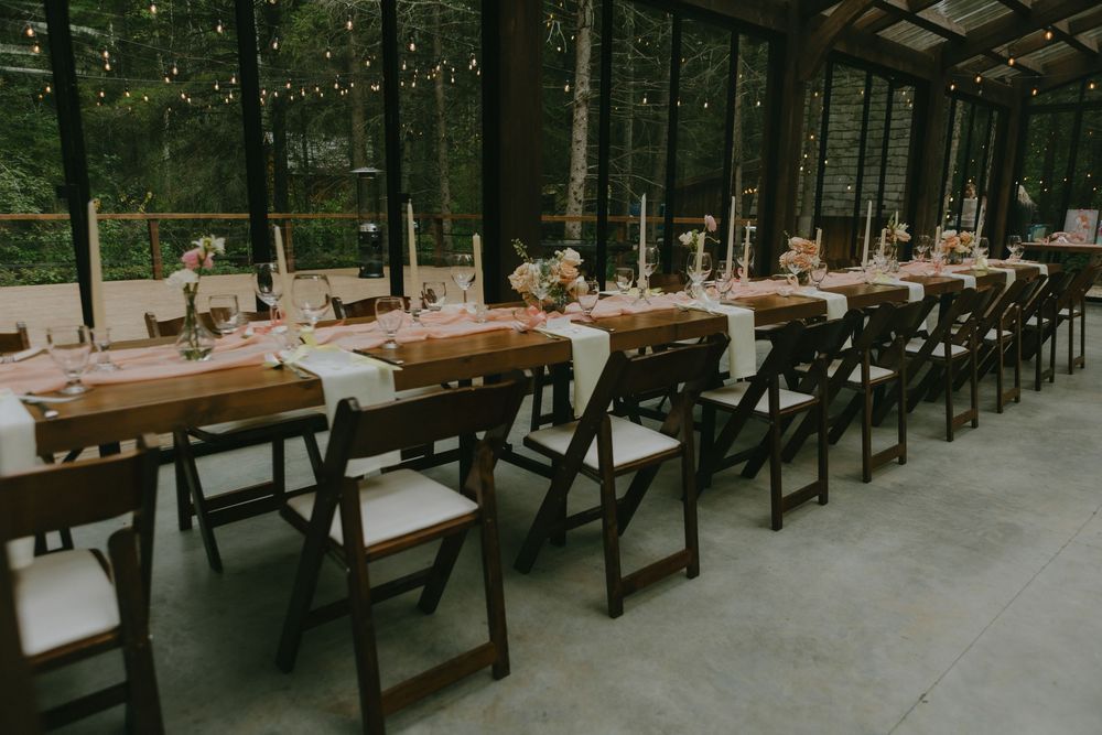 Long wooden dining tables set for a wedding reception under a covered outdoor venue with forest views.