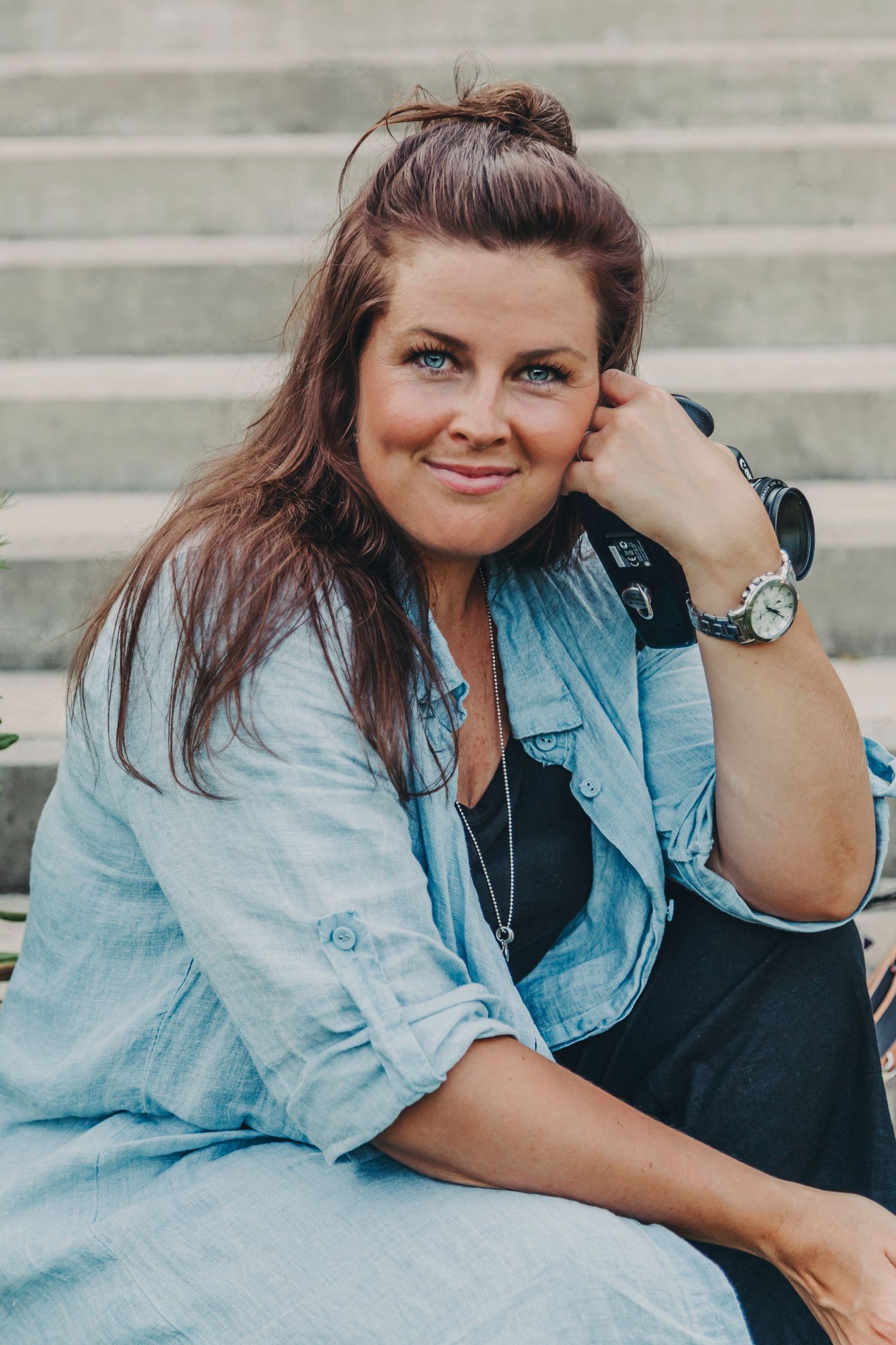 A person in casual denim attire with long brown hair sits outdoors smiling while wearing a watch and earbuds.