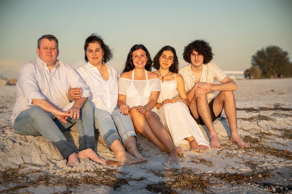 A group sits together on a sandy beach at sunset in light colored clothing, enjoying a relaxed moment.
