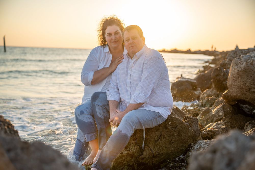 Two people sitting together on coastal rocks during golden hour sunset at a beach with waves in the background.