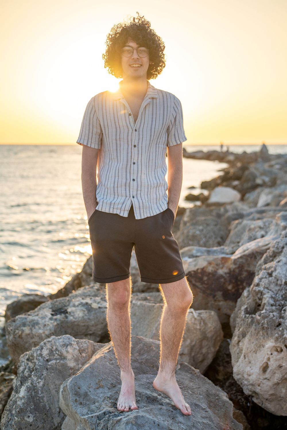 Person in striped shirt and shorts stands barefoot on coastal rocks during golden sunset by the ocean.