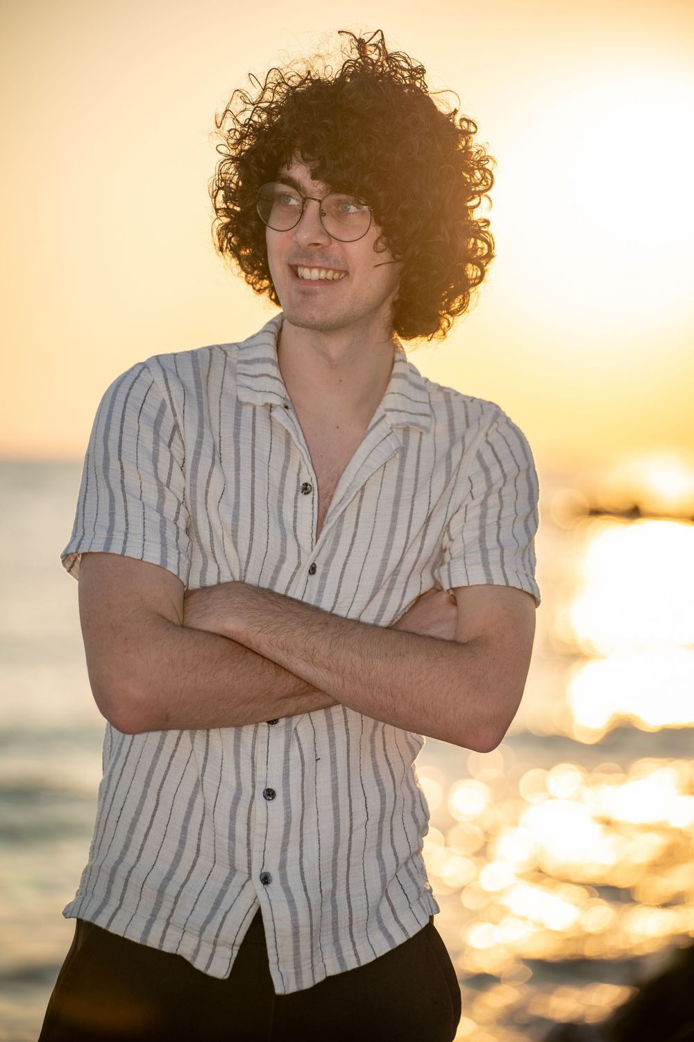 A person with curly hair and glasses wearing a striped shirt stands with arms crossed at sunset by the ocean.