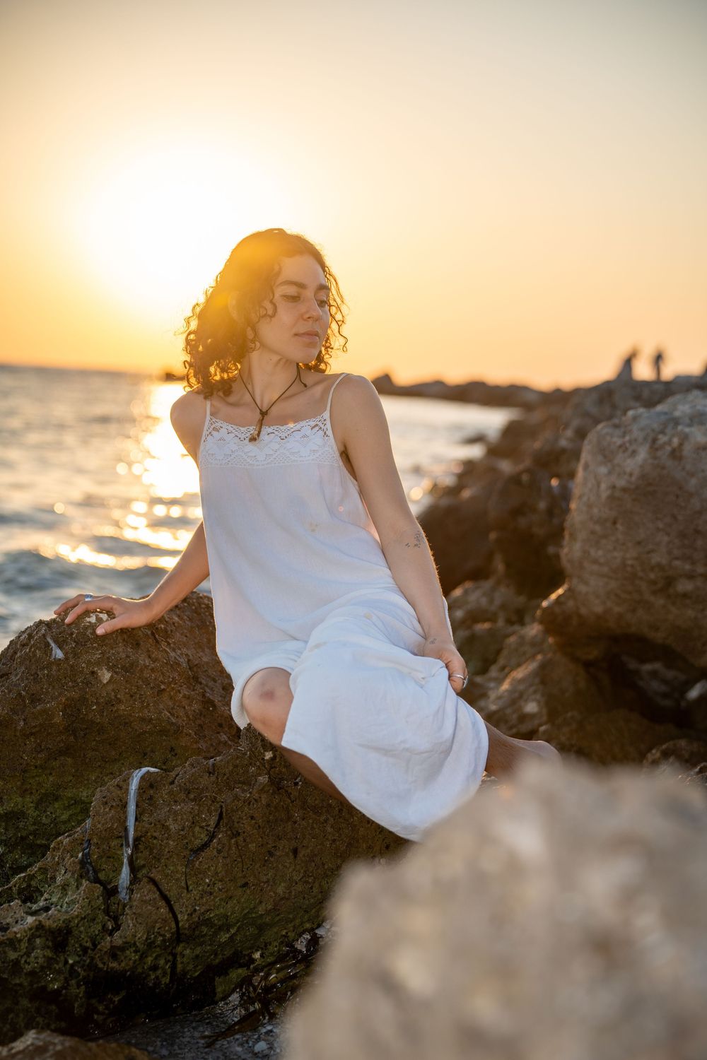 Serene figure in a white dress sits on coastal rocks at sunset as golden light illuminates their curly hair and the ocean waves.