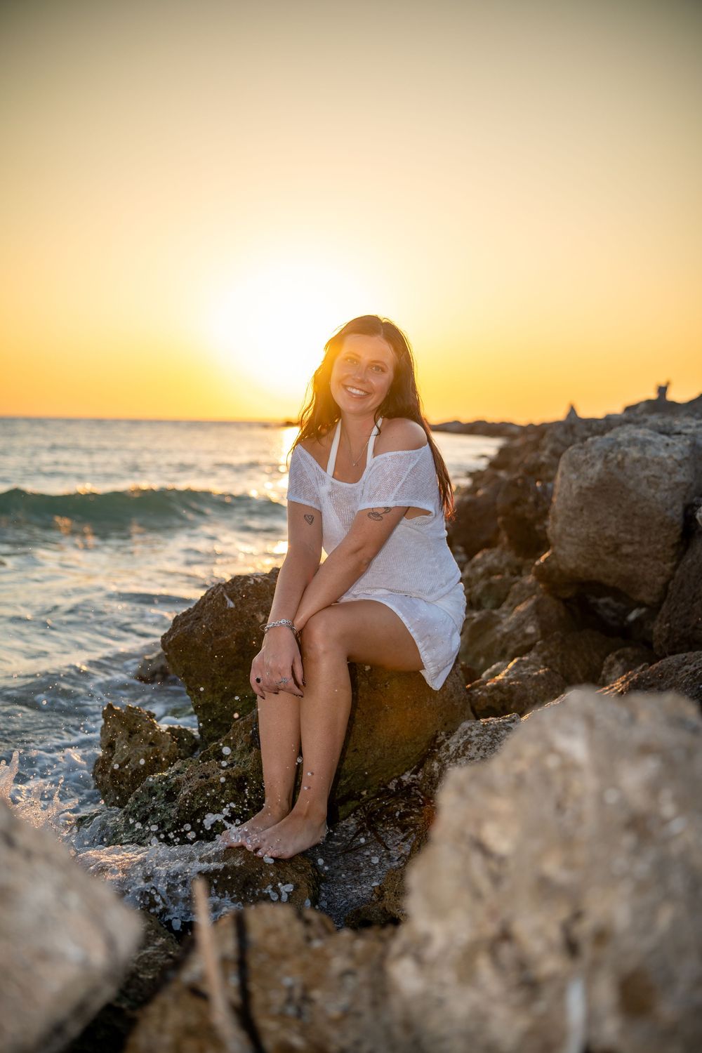 A person in a white dress sits on coastal rocks during a golden sunset over the ocean.