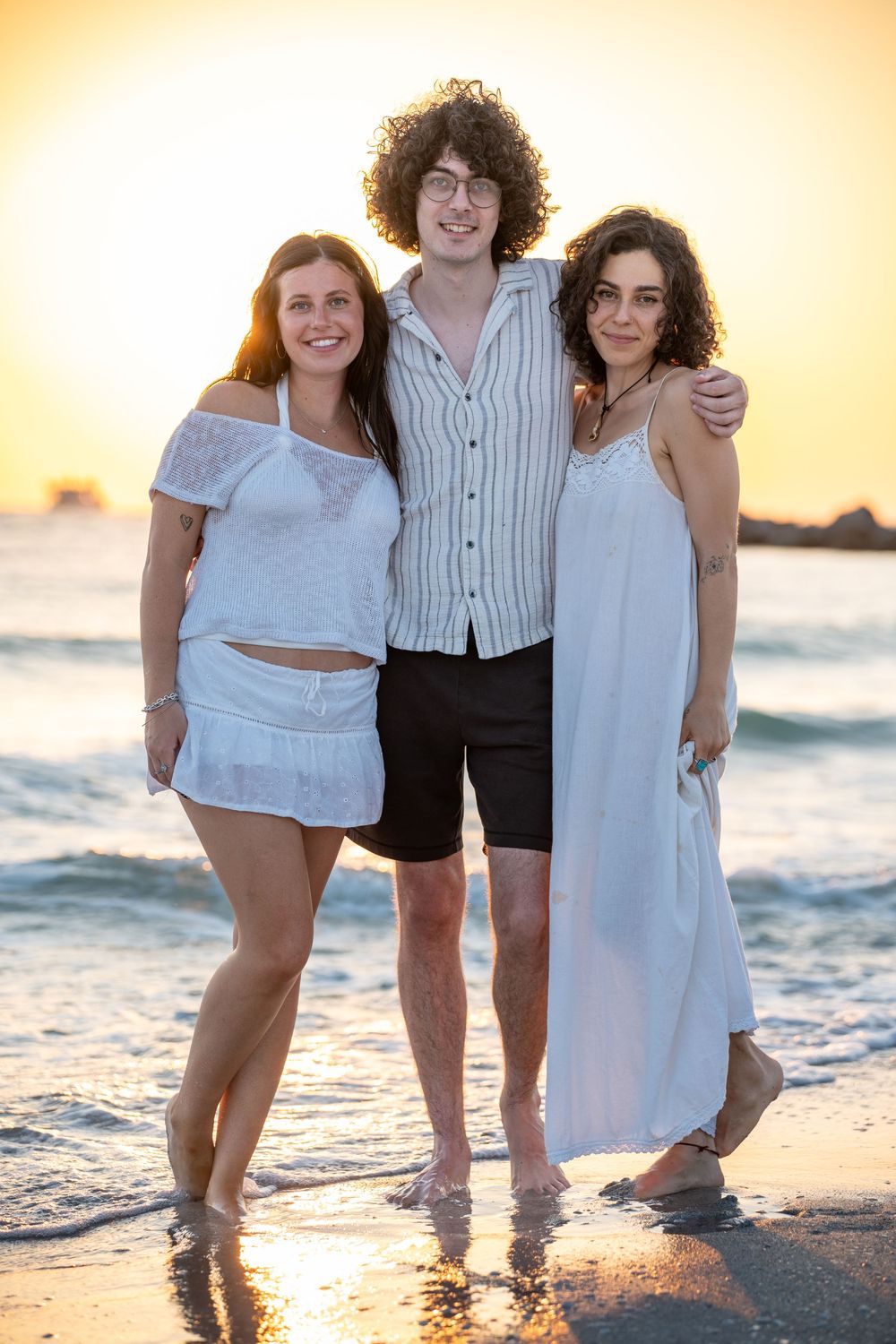 Three people in white summer outfits stand together on a beach at sunset with ocean waves behind them.