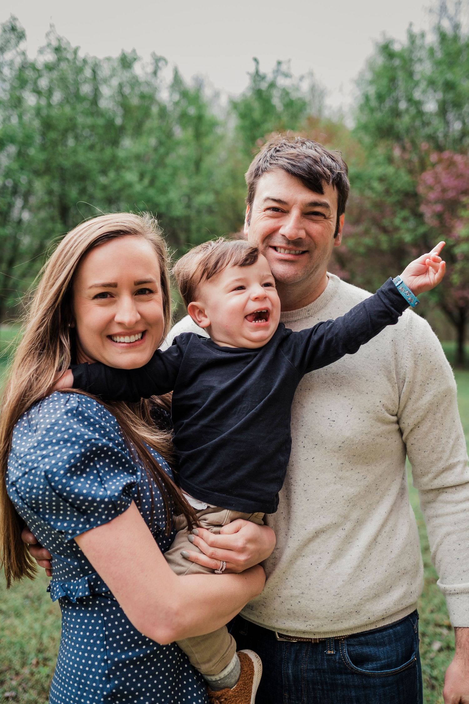 Candid outdoor family portrait of three people with a child pointing and laughing on a cloudy day.