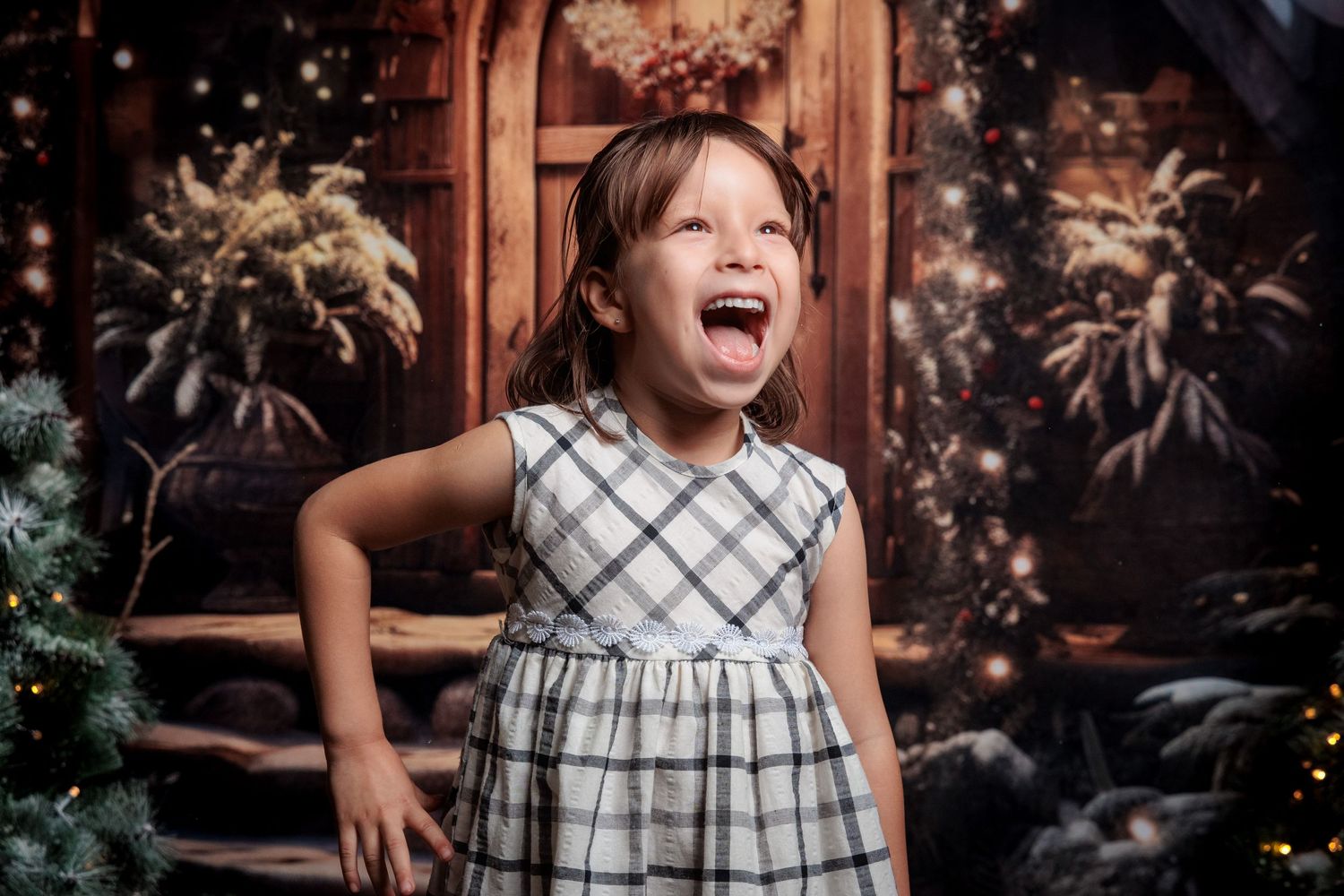 Child wearing a plaid dress poses joyfully against a rustic wooden backdrop with twinkling lights.