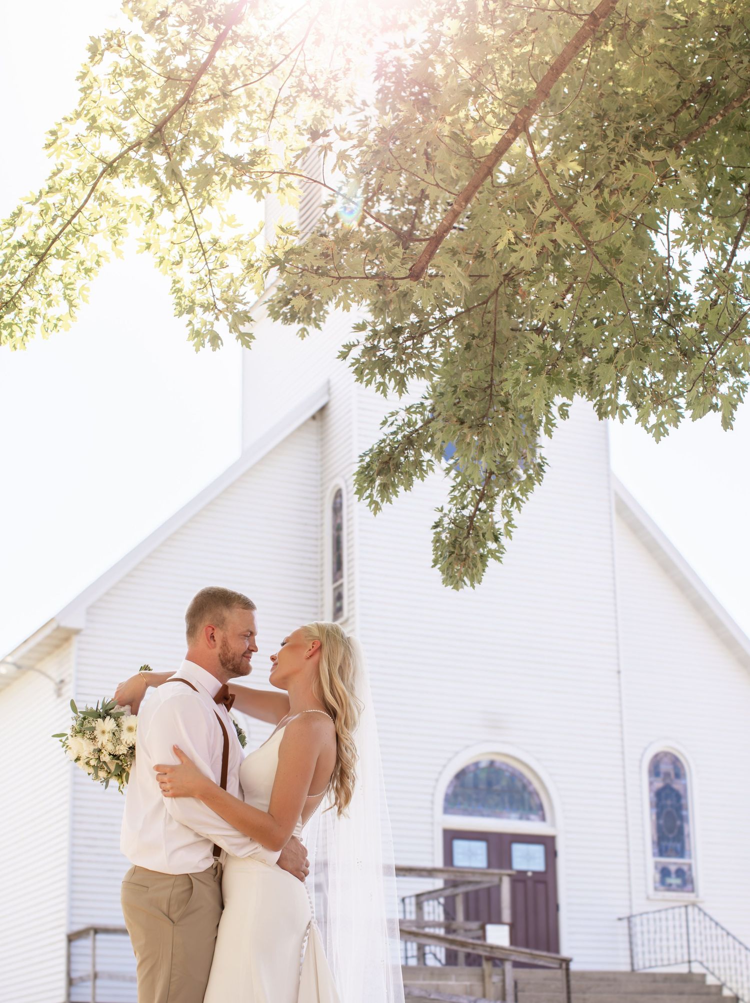Romantic wedding portrait in front of white church with sunlit tree branches overhead.