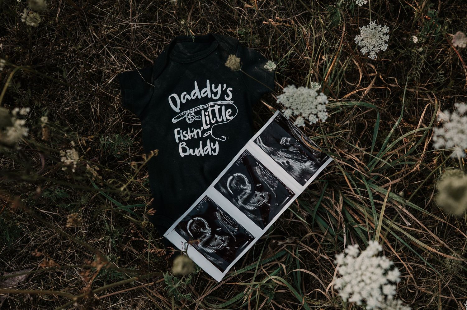 Black baby onesie reading Daddy's Little Fishing Buddy next to ultrasound photos on grass.