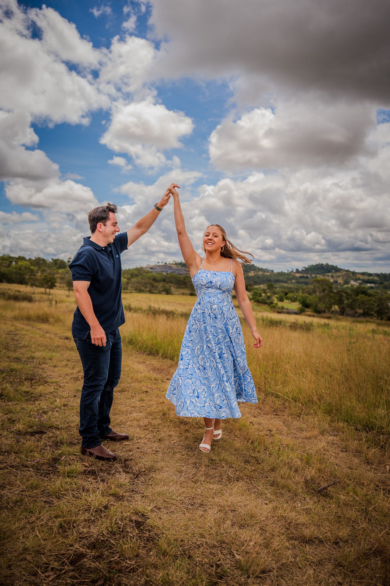 A romantic couple dances joyfully in a sun-drenched field with puffy clouds overhead.