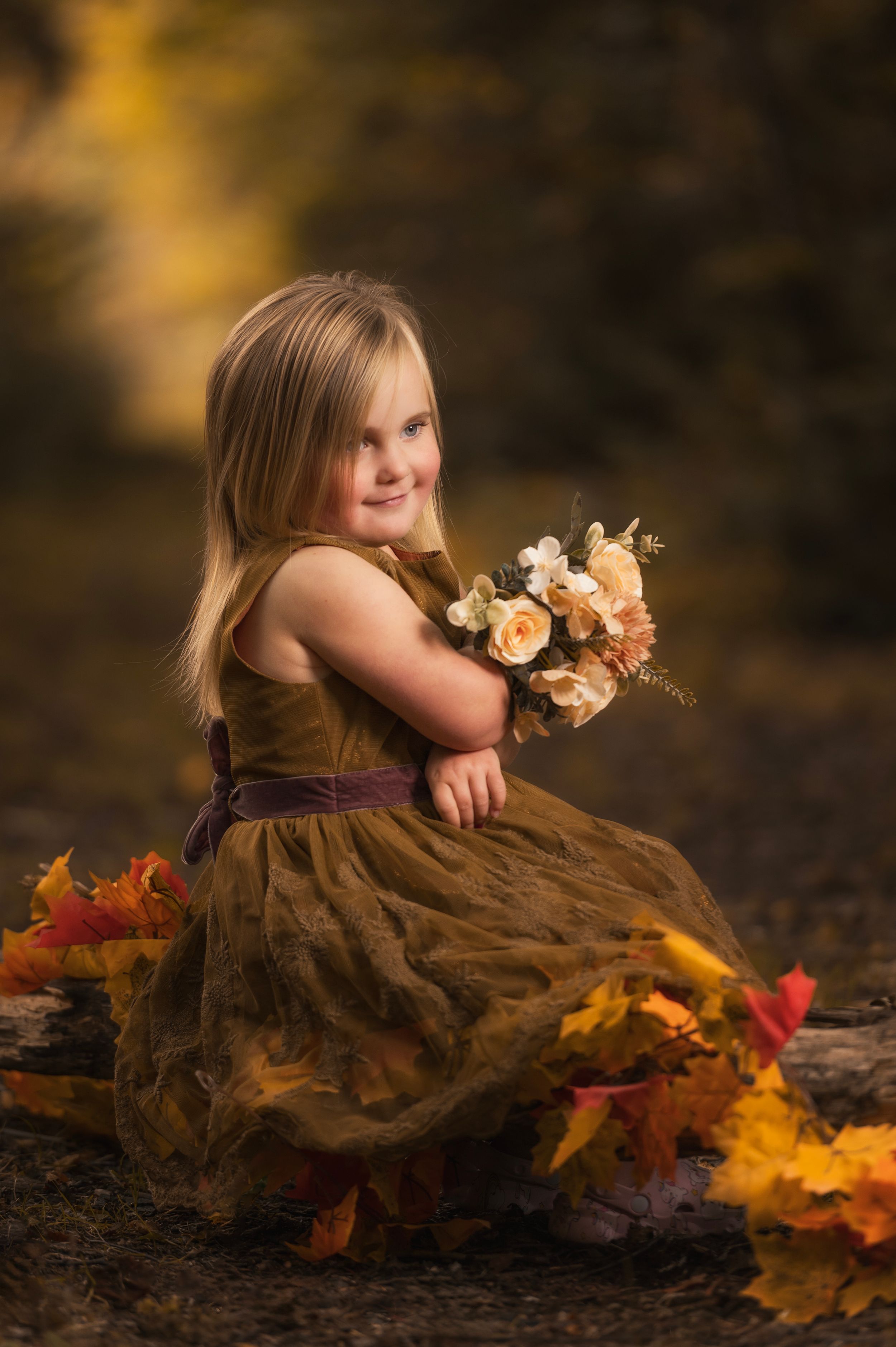 Child in brown dress poses with autumn leaves and peach flowers in warm outdoor setting.