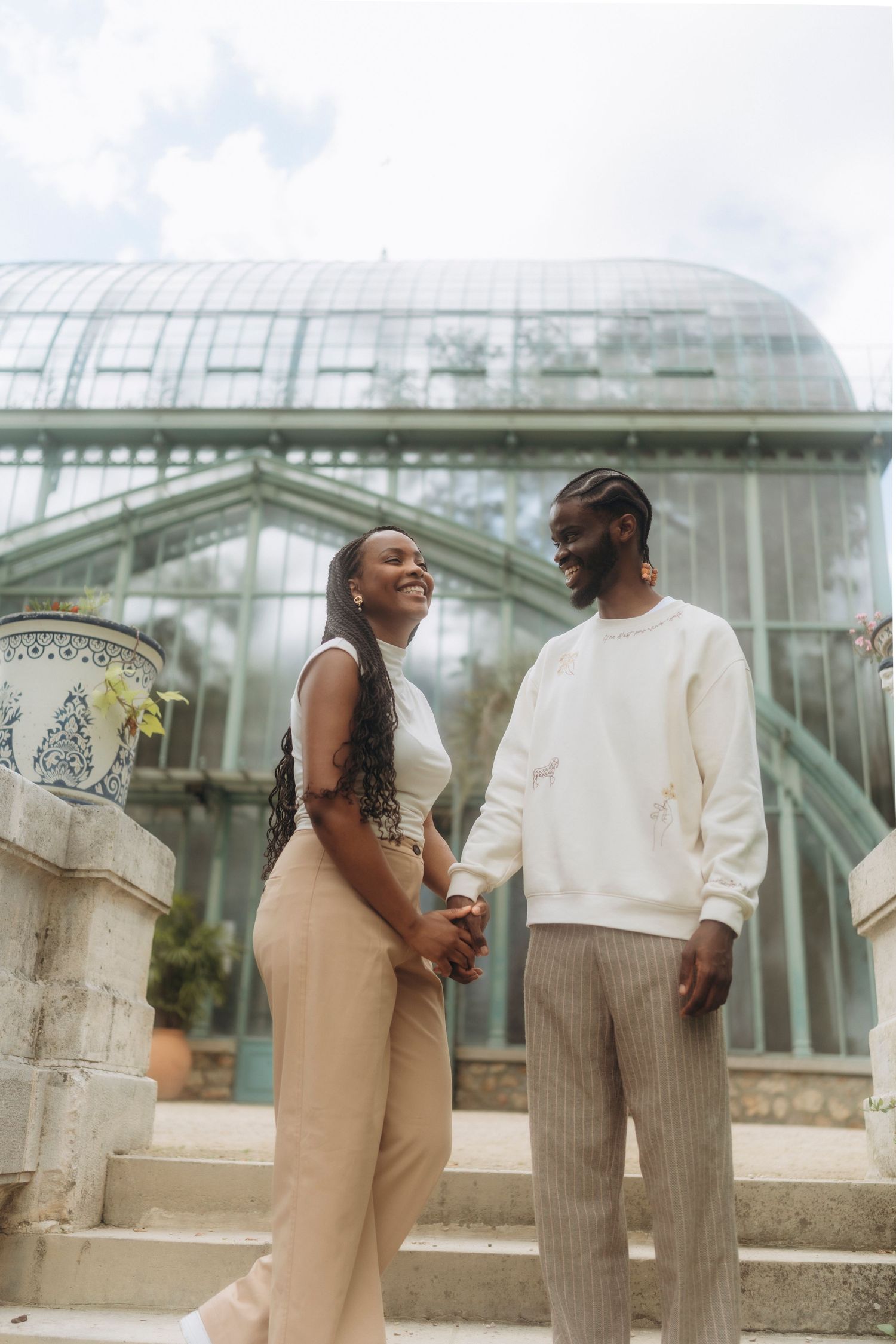 Stylish couple in coordinated neutral outfits pose on steps of a grand conservatory building.