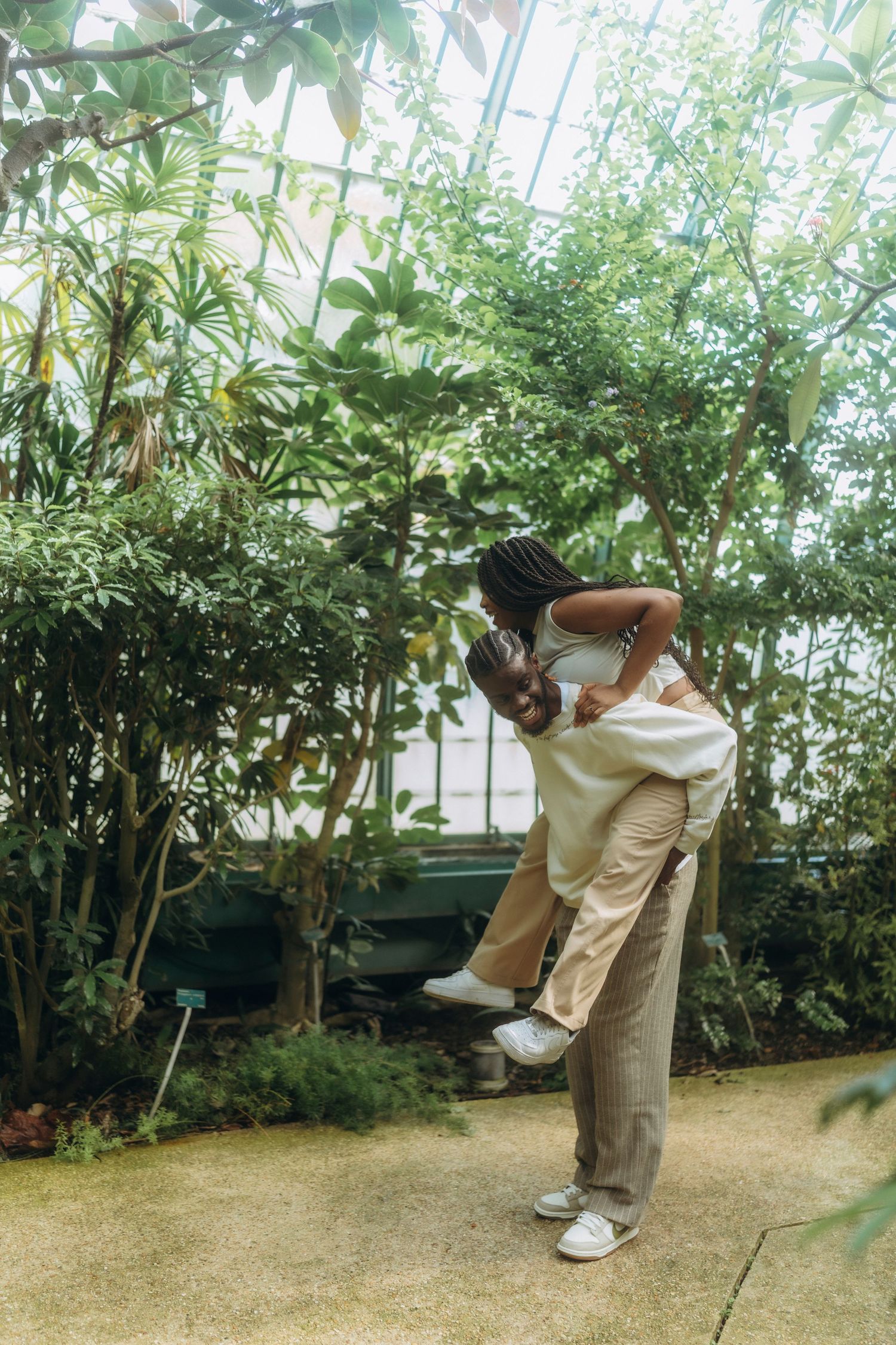 A playful couple shares a lift in a greenhouse filled with tropical plants.