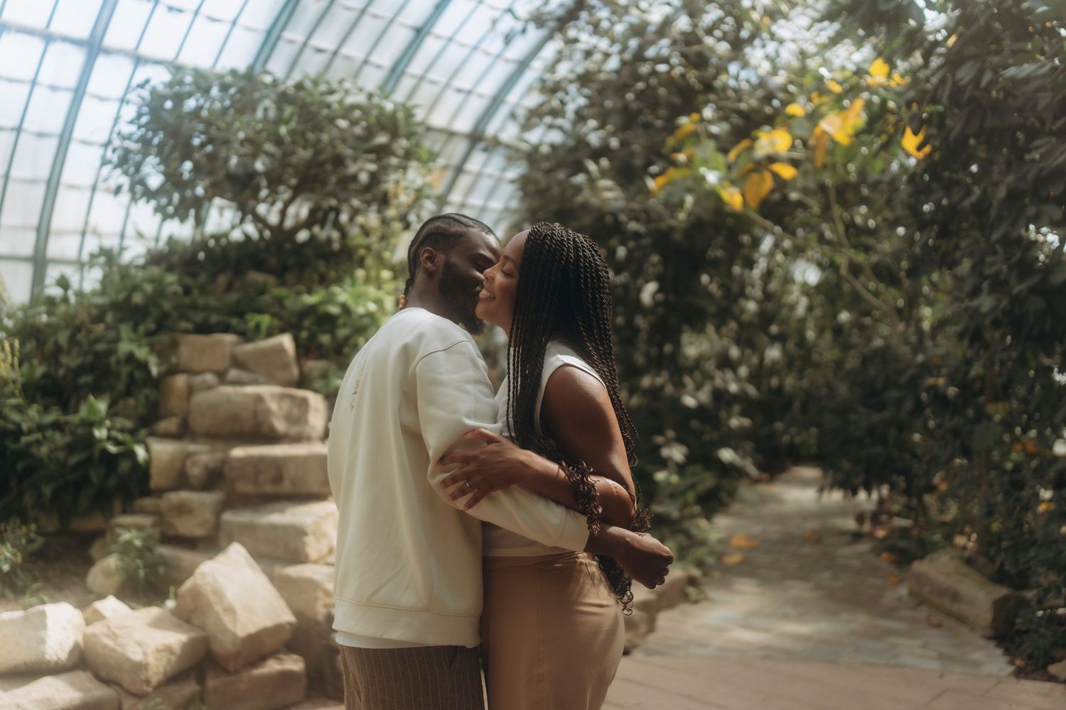 Couple embracing in a sunlit greenhouse surrounded by lush greenery.