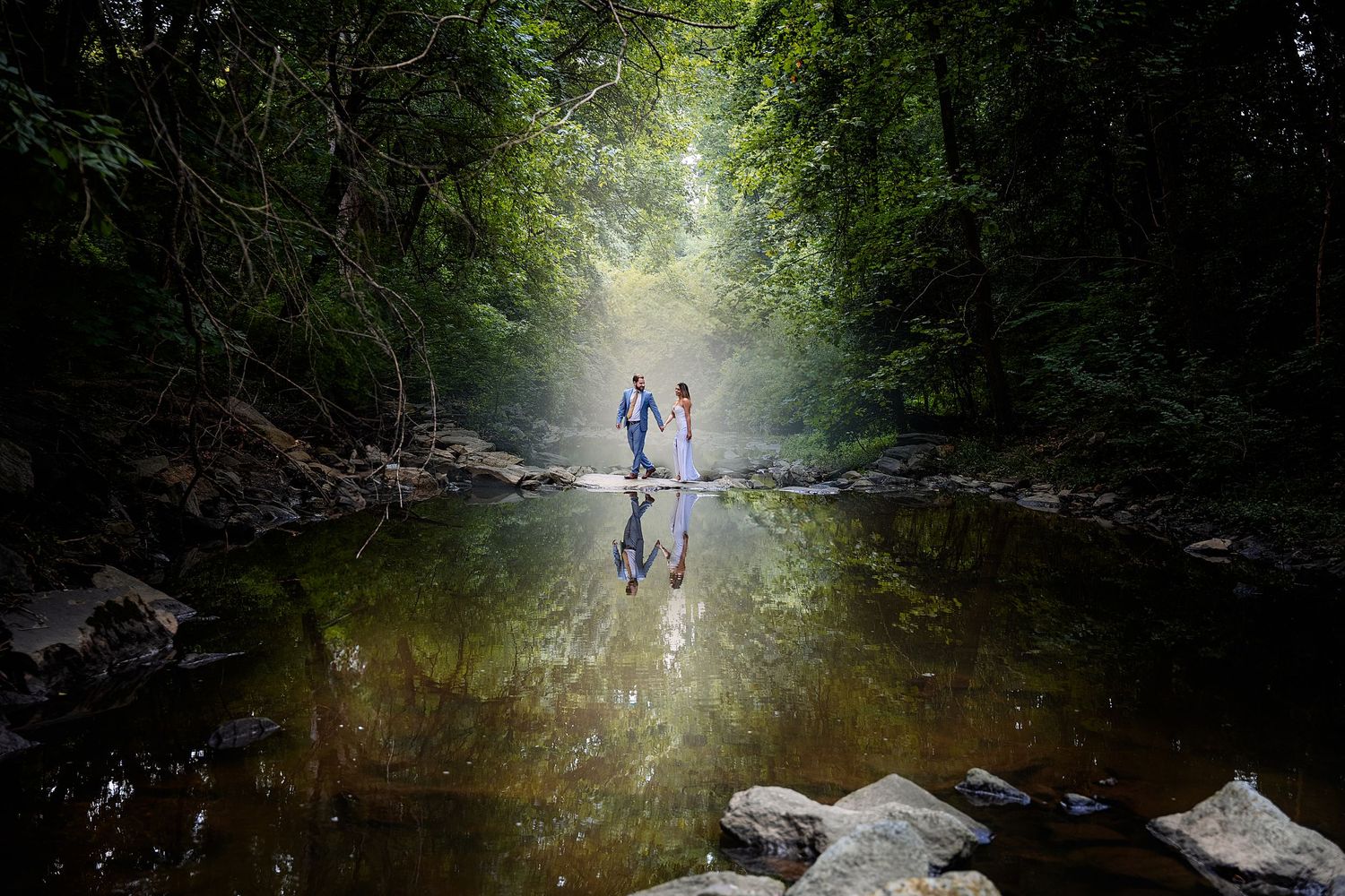 Newlywed couple stands hand in hand on a serene riverbank, surrounded by lush greenery, with their reflections mirrored in the water.
