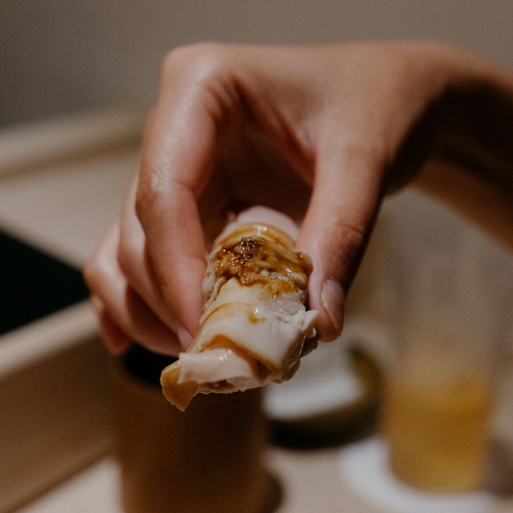 A hand holding a piece of sushi topped with sauce and garnish.