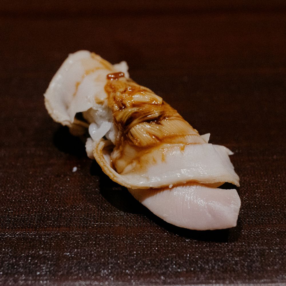 White fish nigiri topped with a golden-brown sauce and glaze on a dark plate.