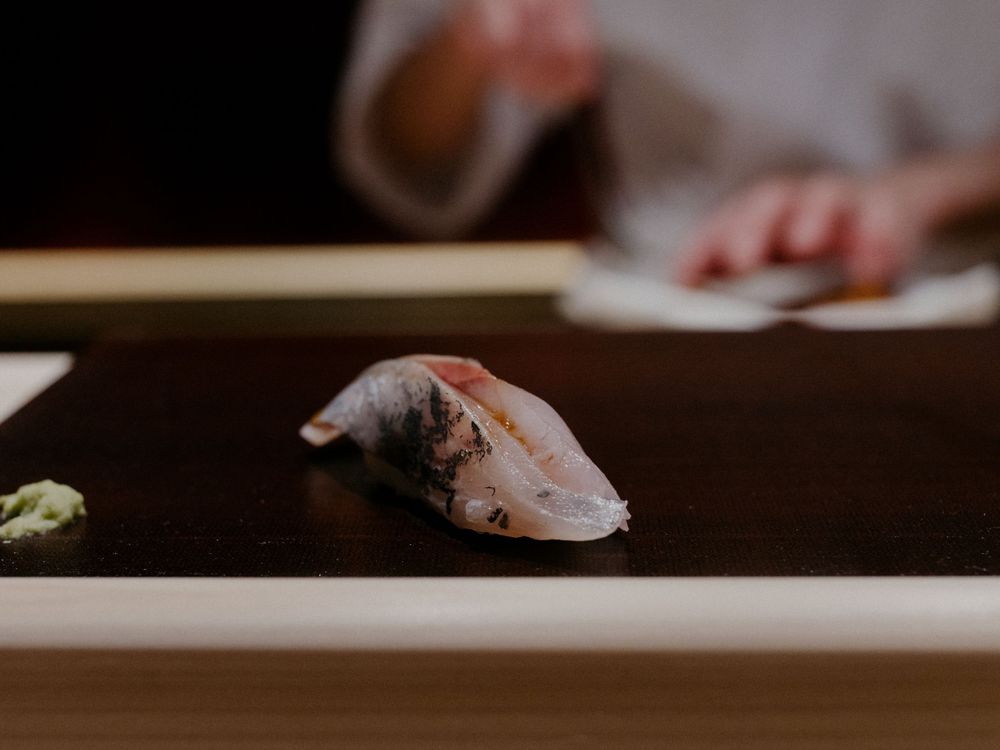 A piece of silver-skinned fish nigiri sushi served on a wooden counter.