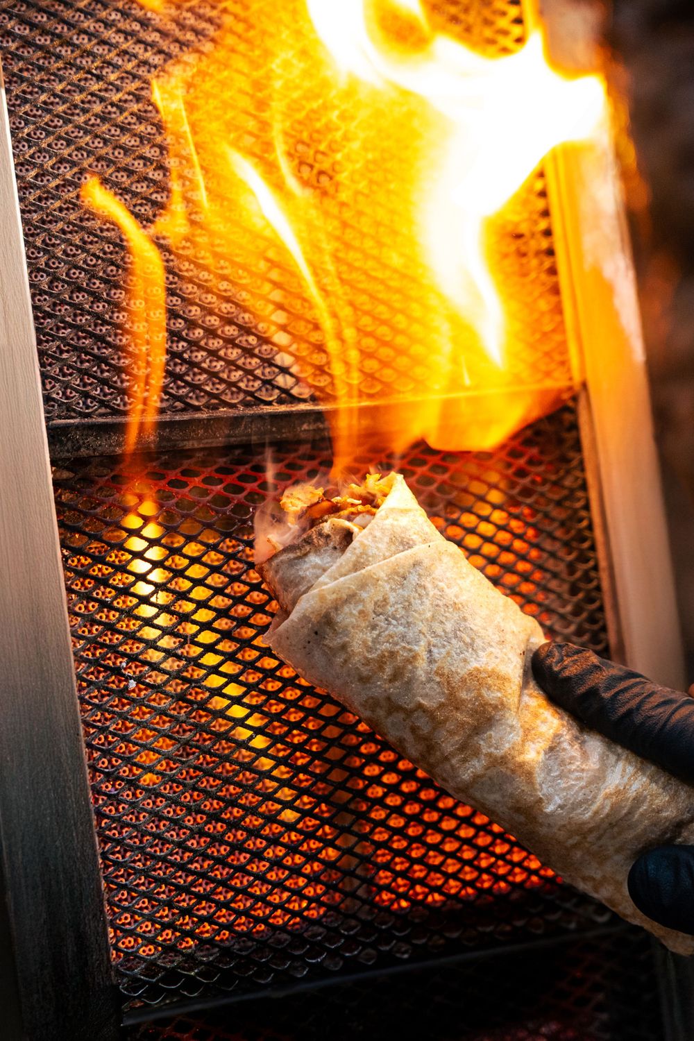 A wrapped burrito being flame-grilled on a metal mesh grate with dramatic fire flare-up. Mtlshutter photography