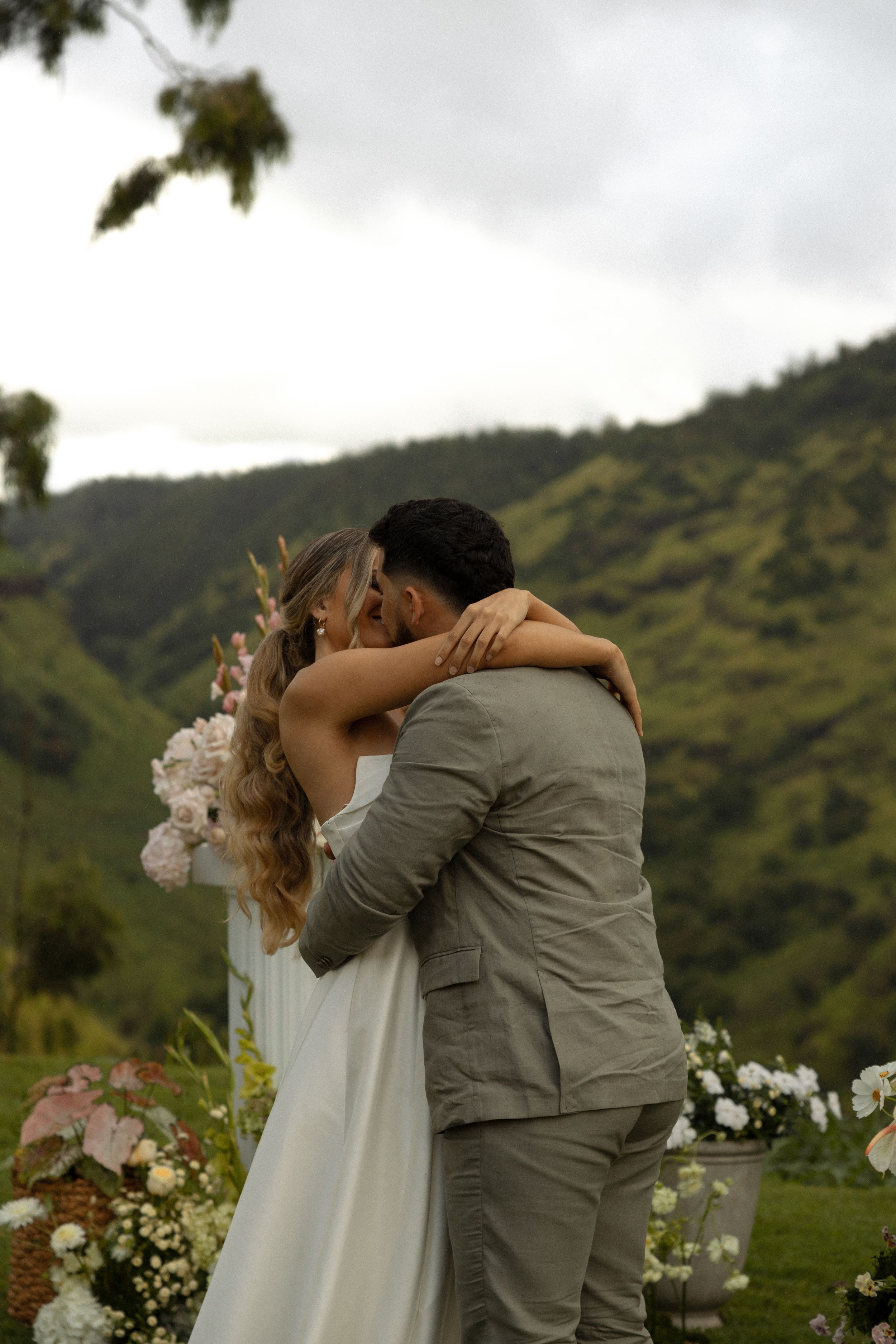 A romantic wedding embrace with a floral bouquet against a lush mountain backdrop in Hawaii.