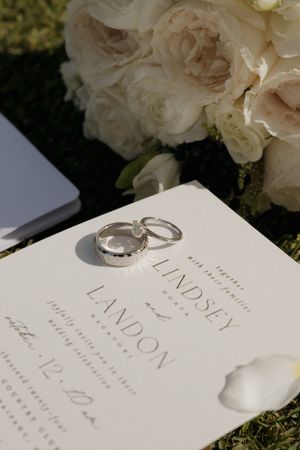 Wedding rings and invitation details displayed with white roses in soft natural light.