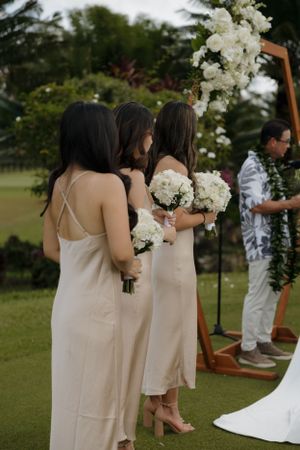 Bridesmaids in champagne-colored slip dresses hold white bouquets during an outdoor wedding ceremony.