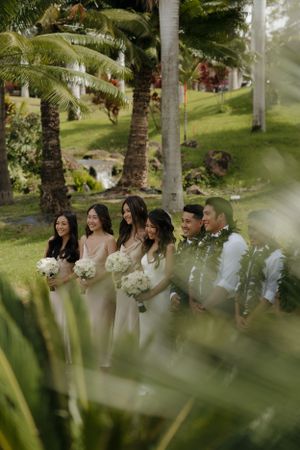 Wedding party in coordinated neutral dresses gather under tropical palm trees at an outdoor venue.