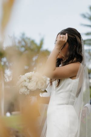 Bride in white dress holds bouquet of white roses while wearing flowing veil outdoors.