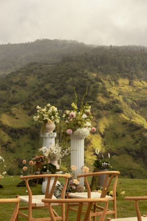 Outdoor wedding ceremony setup with white columns and floral arrangements against green mountains.