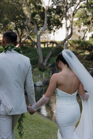 Newlywed couple in white attire holding hands while walking through a garden setting with trees and a pond.