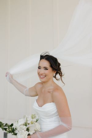 Bride in strapless white wedding gown holds flowing veil while smiling against light background.