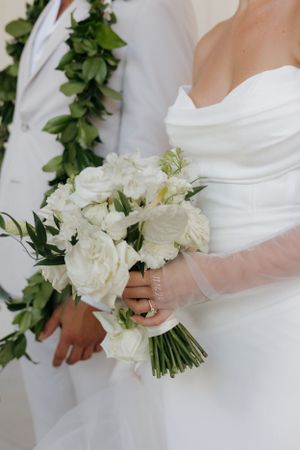 Close-up of elegant white wedding bouquet with roses held by bride in strapless white dress against greenery backdrop.