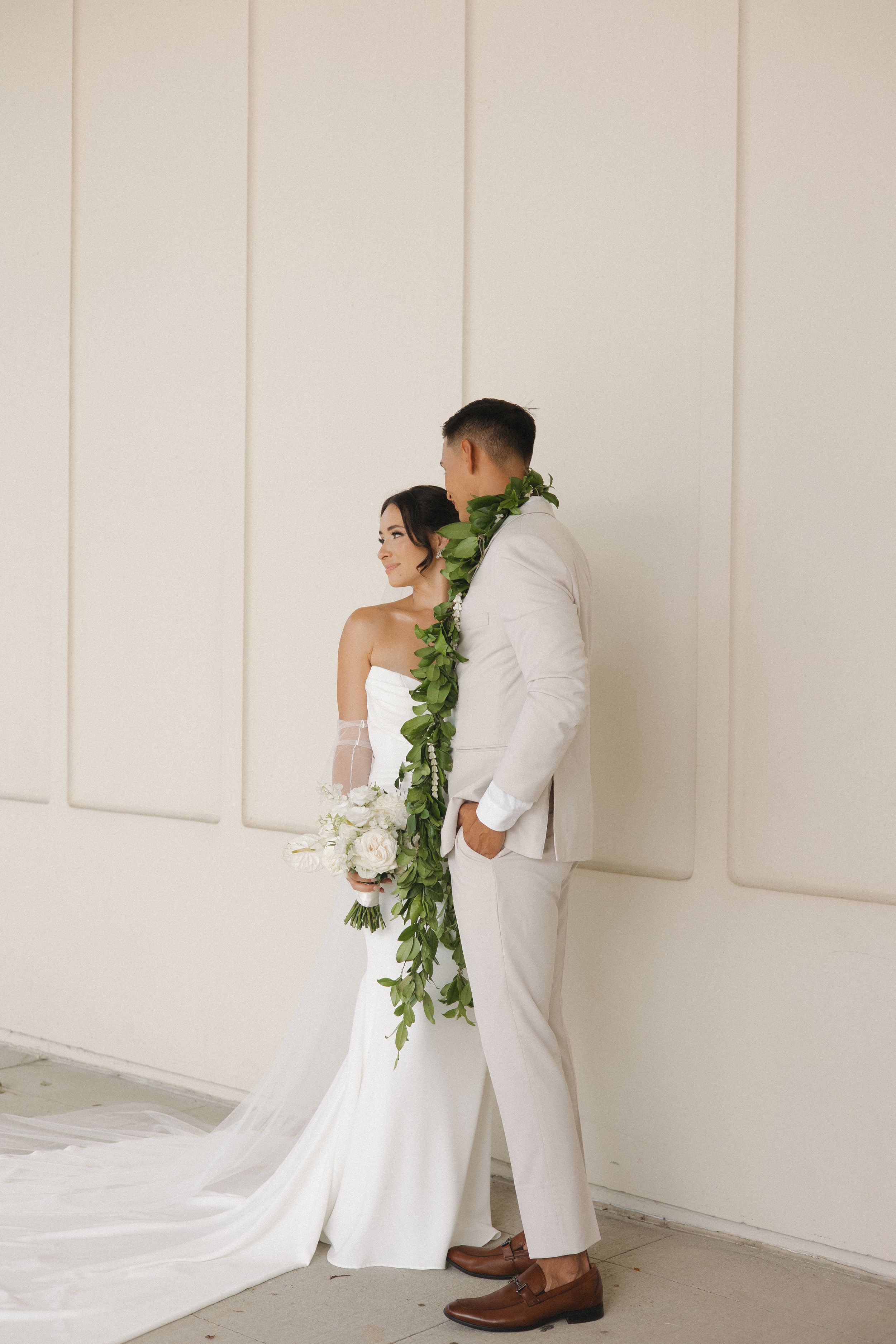 A bride and groom in white attire pose together, adorned with green leaf leis and holding a bouquet.