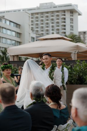 Wedding ceremony at outdoor venue with groom wearing white lei and hotel building visible in background.