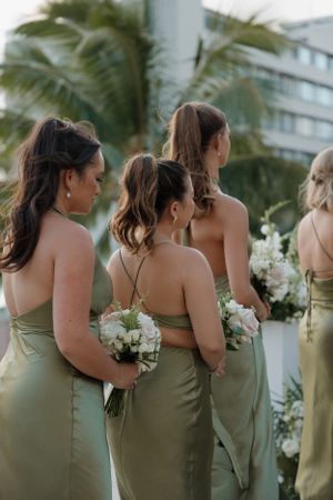 Bridesmaids in sage green dresses hold white bouquets during outdoor beach wedding ceremony with palm trees.