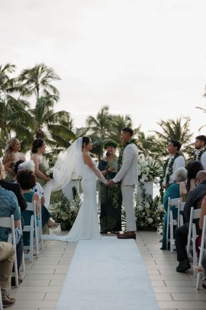 A wedding ceremony takes place on a tropical terrace lined with white chairs and surrounded by palm trees.