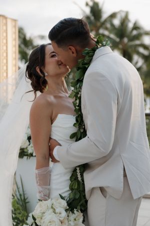 A romantic tropical wedding scene with a bride and groom embracing, with palm trees and white flowers in the background.